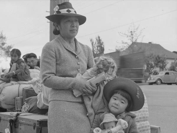 Black and white photograph of a Japanese American woman with a young girl holding a doll in front of a pile of luggage, with another Japanese American family in the background. 