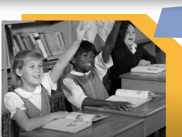 Black and white photograph of three girls sitting in school desks with their hands raised.