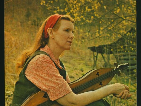 Color photograph of Jean Ritchie holding a mountain dulcimer while sitting on a grassy hill.