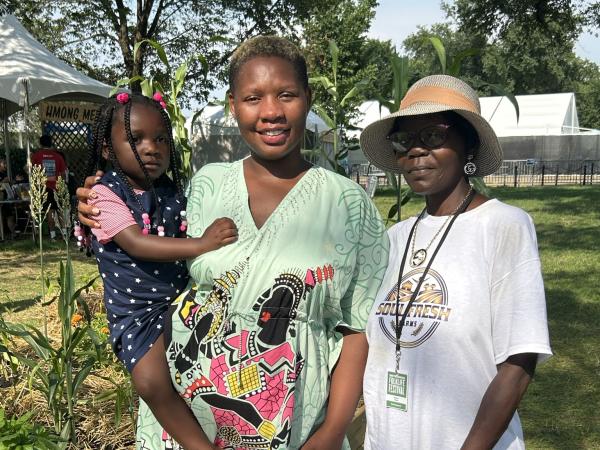 Two African American adult women stand next to a raised garden bed.  The younger woman holds a child and wears a green dress printed with stylized figures.  The woman standing next to her wears a hat and t-shirt with the logo for Soul Fresh Farms.