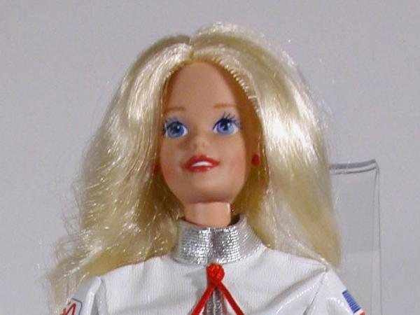 Plastic doll with long blonde hair wearing a white cloth spacesuit with a U.S. flag patch on the arm.