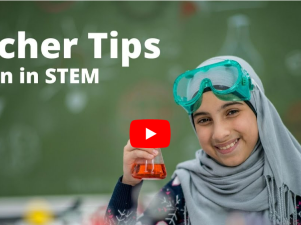 Girl wearing a hijab sits in a classroom and holds up a beaker while smiling