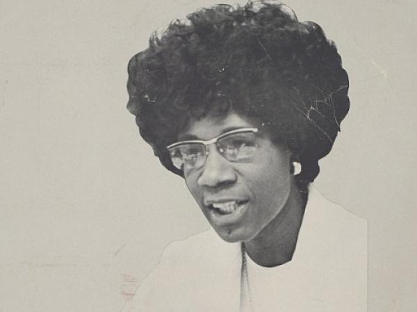 Off-white poster with black and red type. It reads Bring U.S. Together. Vote Chisholm 1972. Unbought and Unbossed. There is a black-and-white image of Shirley Chisholm sitting at a table , speaking and gesturing with her hands, wearing all white.