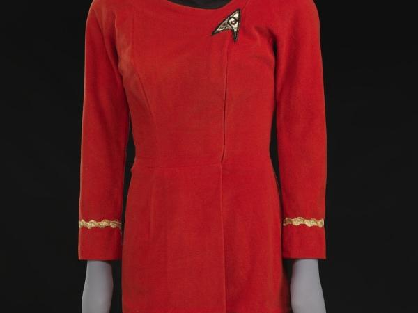 Red dress on a mannequin with gold arm band at sleeves and a wide black collar. On one side of the chest there&#039;s a Starfleet logo patch with gold fabric and black embroidery.