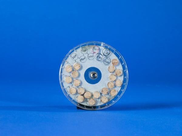 Model of a circular pill dispenser made of plastic, a metal fastener, and a wood dowel