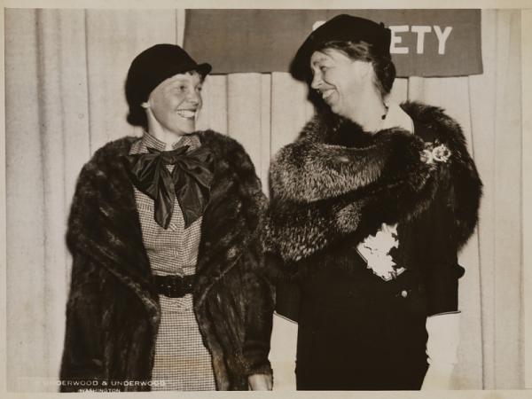 Sepia photo of Amelia Earhart smiling at Eleanor Roosevelt in front of a curtain. They both wear hats, dresses, and fur outerwear.