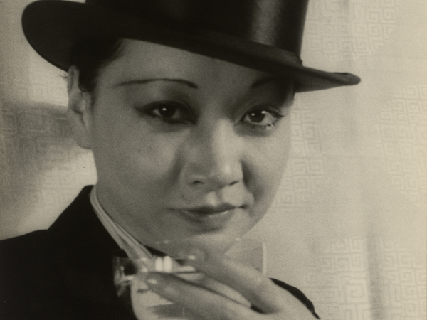 Black and white photo of Anna May Wong wearing a suit and top hat. She arches an eyebrow and holds a glass to the camera in a toast.