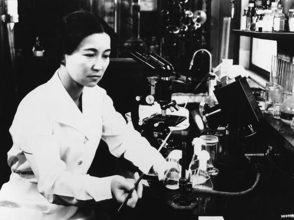 Ruby Hirose sits at a lab station. She wears a white lab coat and holds a pipette.