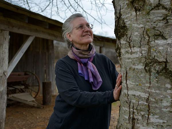 Photograph of a woman with gray hair, wearing glasses, a purple-toned neck scarf, and dark clothing looking at a large tree with both of her hands on the trunk.  A wood shelter with the wheel and tongue of a horse-drawn wagon visible is in the background. 