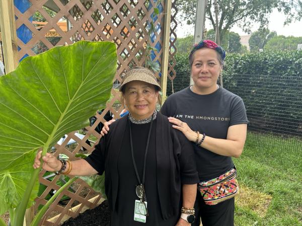 Two Asian American women wearing dark clothing stand next to a banana plant in front of a lattice wall. The woman in front is older and shorter than her daughter standing behind her who has her hands on her mother’s shoulders.