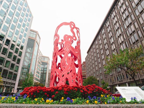 Photo of Marker, a tall cylindrical metal sculpture depicting pink intersecting threads. It stands above a bed of colorful flowers.