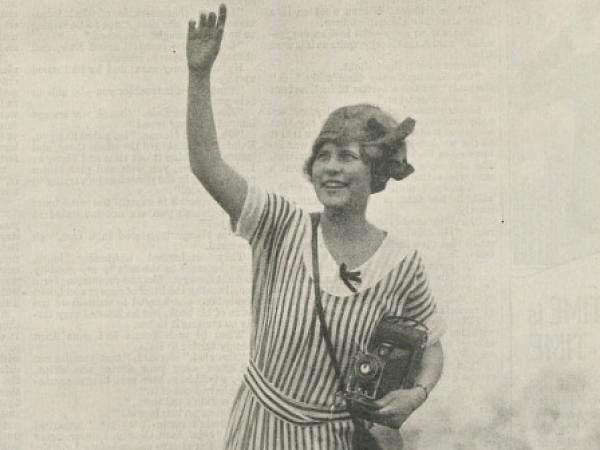 Advertisement with a black and white photograph of a young woman wearing a striped dress. She is holding a camera in her left hand as she stands on the bottom beam of wood fence and waves her right hand.