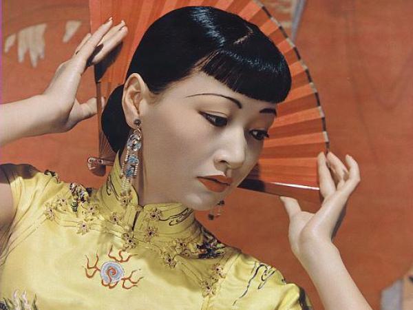 Anna May Wong poses, holding a fan behind her head. She wears a yellow dress with dragons emblazoned on it.