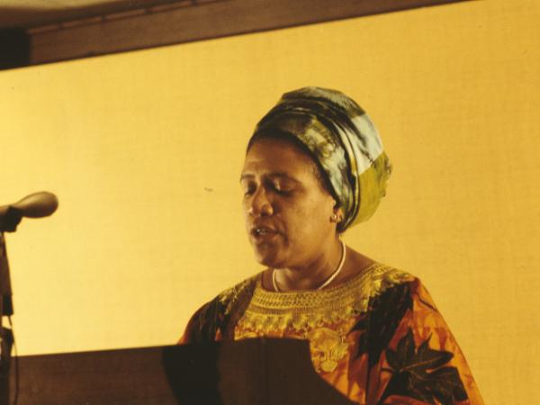 Candid color photo of Audre Lorde from the shoulders up standing at a microphone and podium.   