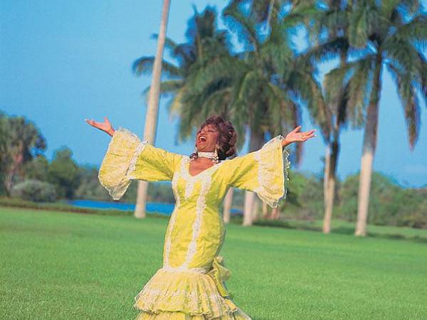 Celia Cruz stands outside on manicured grass with her arm stretched wide. She wears a bright yellow bata cubana (Cuban rumba dress) with wide bell sleeves. Her mouth is open, as if she is singing or shouting. There are palm trees in the background.