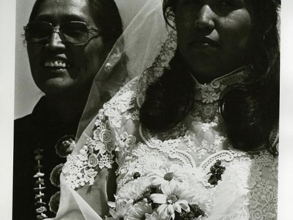 Photo of Thelma Begay Callan [Diné (Navajo)] dressed in her non-traditional wedding dress and holding flowers with her mother, Mitzi [Diné (Navajo)], standing behind her wearing traditional clothing