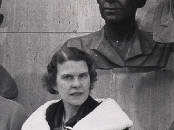 Black and white photograph of a woman standing in front of her own portrait bust wearing a dark coat with a white collar.