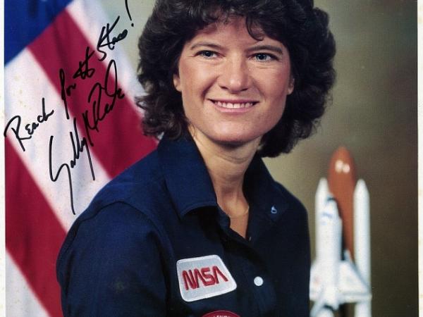 Sally Ride headshot with American flag and a model of a space ship behind her. The photo is signed: Reach for the stars! Sally Ride