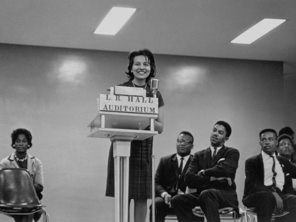 A black-and-white image of Diane Nash at the podium with men and a woman sitting on either side of her in chairs. Nash stands in the center of the image behind the podium labeled [L.R. HALL/ AUDITORIUM] speaking into the microphone.  