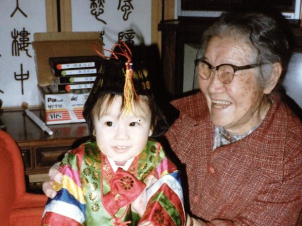 A color photo of Sung Hark (Lee) Kang sitting on a red carpet holding up Nicole as a toddler.    