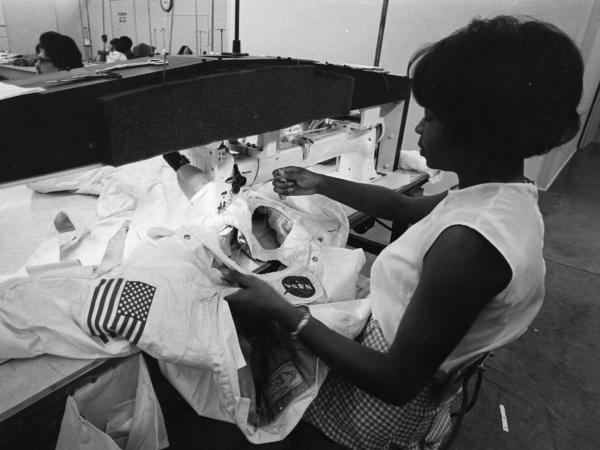 Black and white photograph of African American woman sitting at a long-arm sewing machine working on the iconic white outer layer of an Apollo spacesuit.