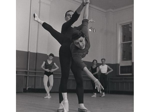 Ballerina Maria Tallchief rehearses a dance on pointe, extending one leg into the air. A male dancer holds her hand to support her movement. Other dancers watch in the background.