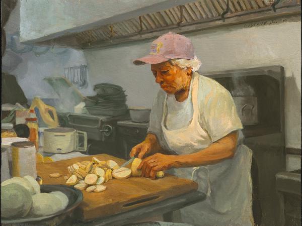 Woman cutting a yellow squash in a restaurant kitchen. Steam rises from a sink behind her. She wears a baseball cap.