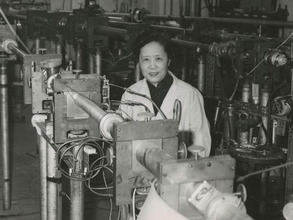 Chien-Shiung Wu, smiling at the camera and wearing a lab coat, posing among unidentified apparatus