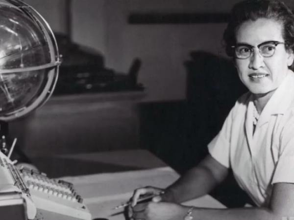 Katherine Johnson looks at the camera. She sits at a desk with papers for calculations and a large globe.