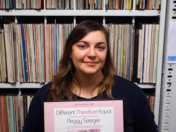 Meredith Holmgren holds a record that says "Different Therefore Equal / Peggy Sawyer. "She stands in front of a shelf full of records. 