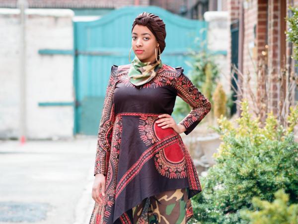 Woman wearing a fashionable ensemble of tunic, pants, headwrap, and neck scarf. The pants and neckscarf are made of a camoflauge print fabric. The tunic is made of an African wax-print fabric.