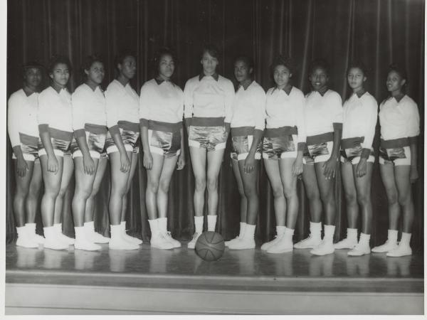 A black-and-white photograph of eleven members of a women&#039;s basketball team standing in front of a curtain. The tallest woman stands in the middle, with the individuals being shorter as they stand further from the center. A basketball sits at their feet.