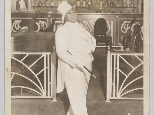 A silver gelatin print depicting a black-and-white image of entertainer Gladys Bentley. She is wearing a white tuxedo, top hat, and she is holding a cane under her proper right arm.