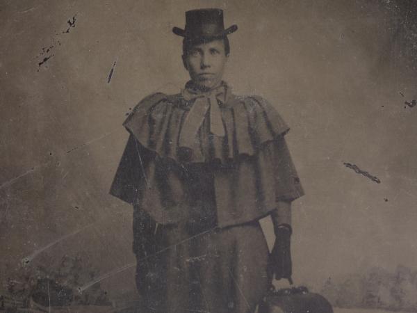 A tintype occupational portrait of a woman, likely a doctor or possibly a nurse or midwife, carrying a medical bag. The woman is standing tall at the center of the image, looking directly at the camera. She carries a medical bag in her proper left hand.