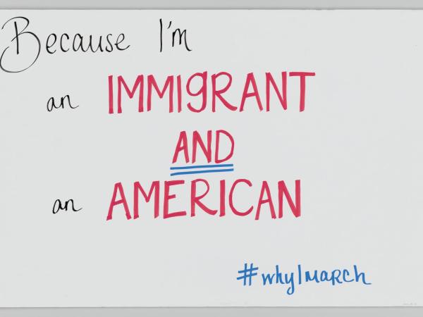 The poster has handwritten black and red text which reads &quot;Because I’m an IMMIGRANT AND an AMERICAN.&quot;