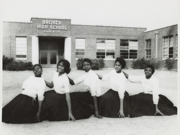 A black-and-white photograph of five young Black women in light colored tops and dark skirts seated in a line in front of Breisch High School