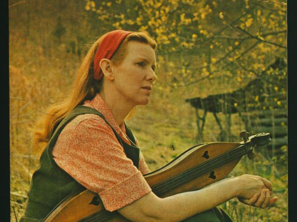 Jean Ritchie sits in a field, holding a plucked dulcimer. She stares off into the distance, thoughtful.