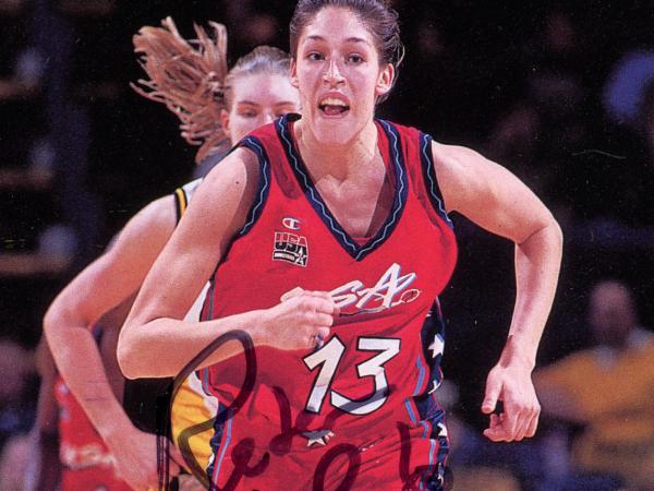 Rebecca Lobo runs towards the camera, as an opposing player runs behind her. She wears a red and black Team USA jersey.