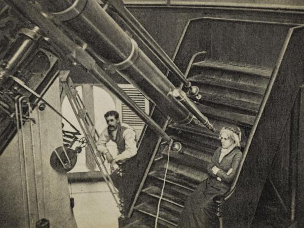 This postcard shows a woman sitting on the observing chair in front of a large refracting telescope