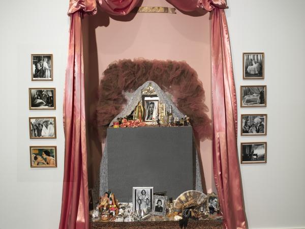 Draped sumptuous pink fabric frames rows of black-and-white photos and an arrangement of found objects including a fan, bracelets, painted fruit, dried flowers, and lace, all set in a niche.