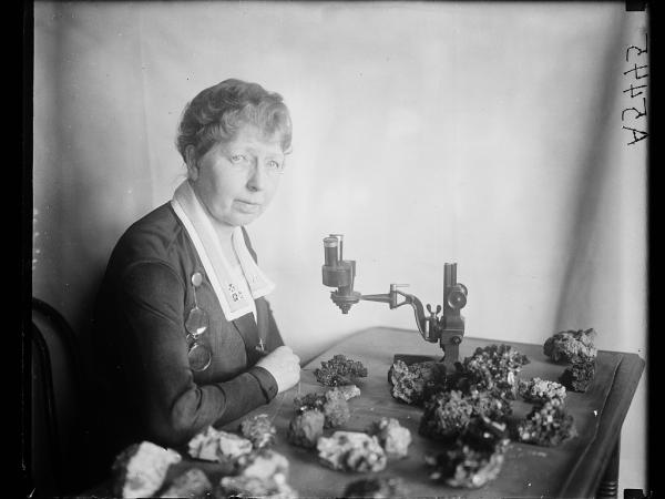 Black and white photo of Margaret W. Moodey sits at a desk with a microscope and gemstones