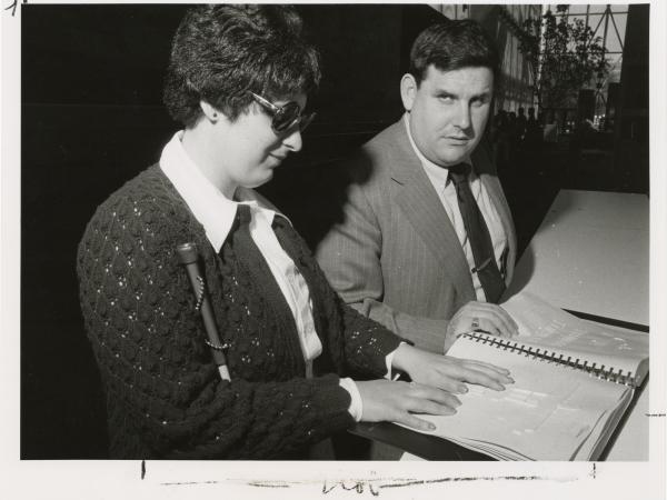 Renee, a blind woman, examines raised line drawings of the National Air and Space Museum as another staff member watches her. Renee wears dark glasses and carries a cane.