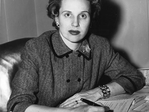 Wynne Sharples Ballinger sits at her desk, which is covered in documents. She holds a pen. She stares straight at the camera and does not smile.