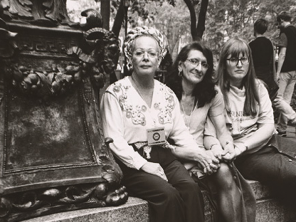 Sylvia Rivera, Christina Hayworth and Julia Murray sit on a ledge in a park in New York City. A sign near them reads: Respect Trans People/Men!