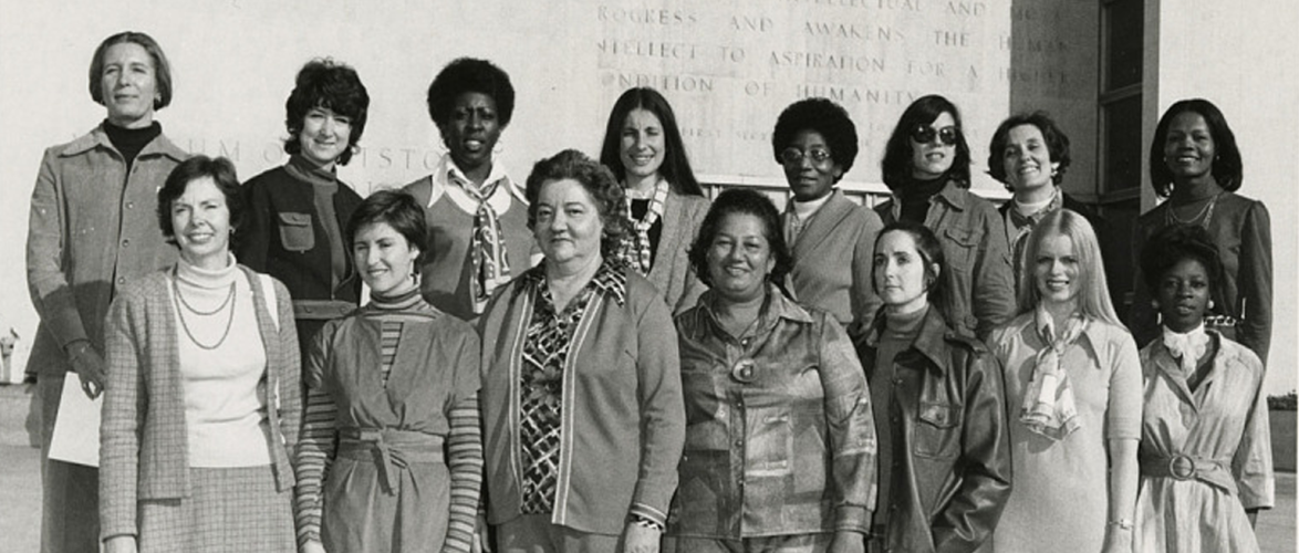 Black and white photo of fifteen women, with a variety of skin tones, standing in two rows