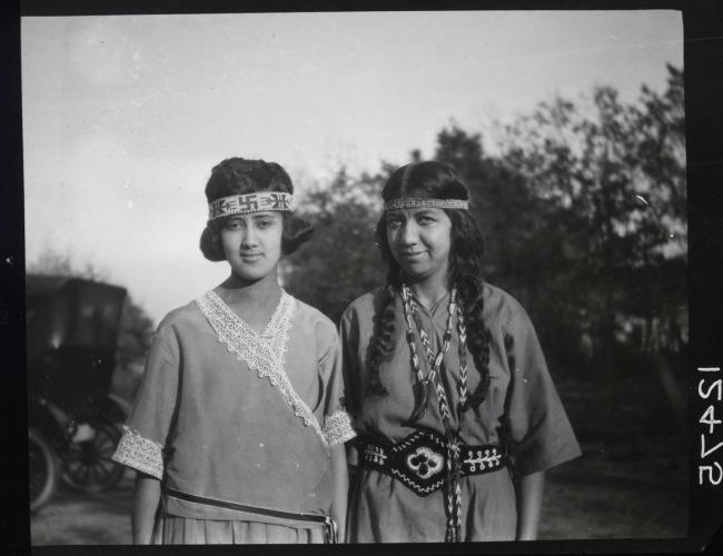 Jane Harmon and Gladys Tantaquidgeon stand next to each other and look at the camera. They wear loose 1920s era clothing with Indigenous accents, including belts and headbands 