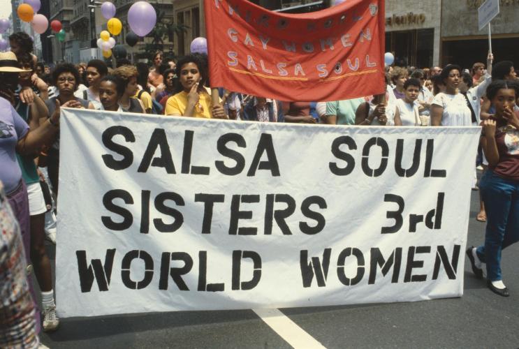 A color photographic slide of marchers in the New York City Gay Pride Parade holding a white banner with the words SALSA SOUL SISTERS 3RD WORLD WOMEN printed in black