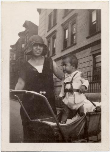 Clotilde Arias stands on a city street, smiling and holding up her son, who is standing in a baby carriage