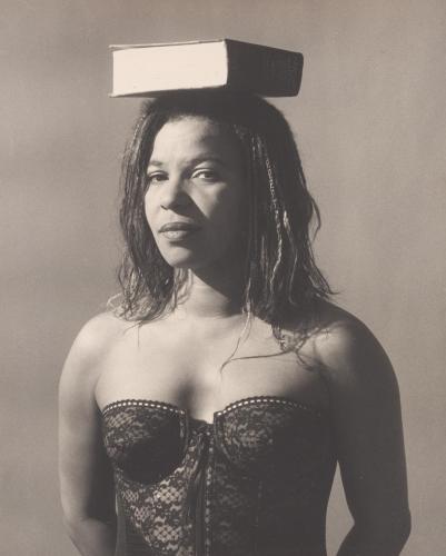 Sepia photo of Ntozake Shange balancing a book on her head. She wears a corset and her hair is in thin braids