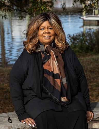 An African American woman wearing dark clothing and a scarf colored in earth tones sits on a stone bench with a river in the background.